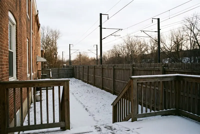 Tall solid wood privacy fence along the rear of a narrow back yard, with a commuter rail line and catenary beyond