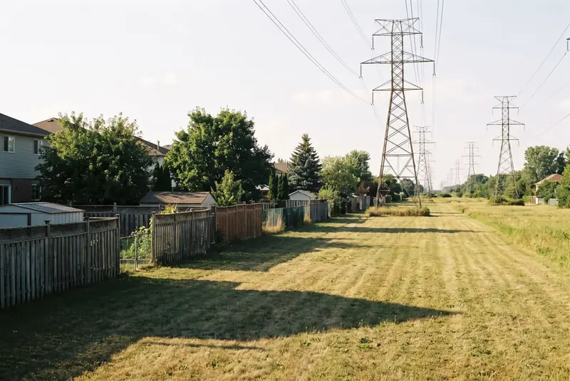 Open-mown utility corridor with steel transmission towers behind a row of suburban backyards with fences stopping short