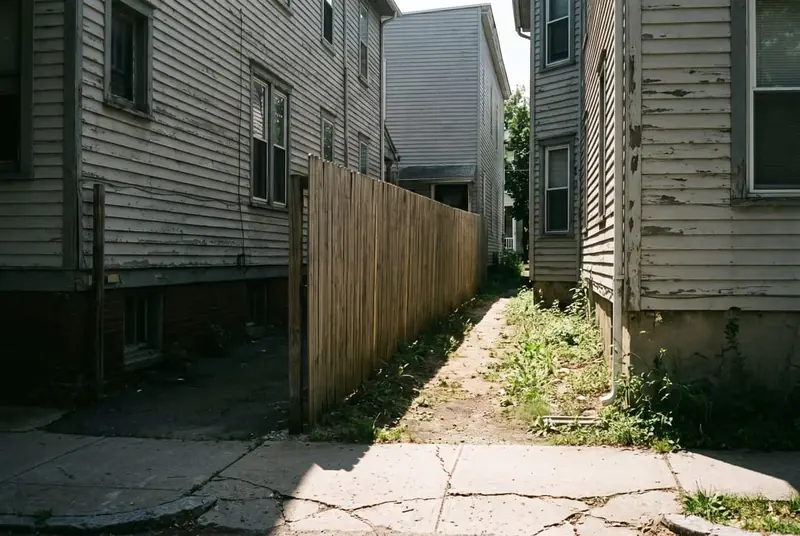 Narrow side yard between two older houses with a tall solid board fence close to a neighbor's side windows