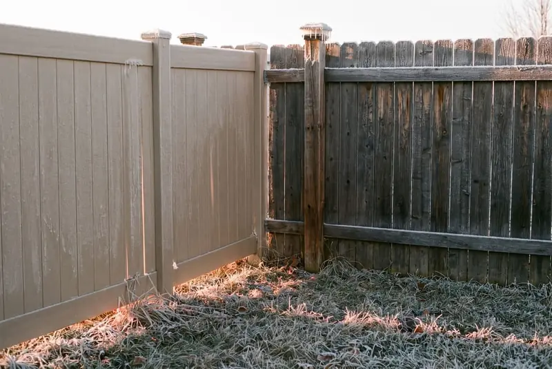 Tan vinyl privacy fence and a stained cedar fence side by side along a frosty backyard lawn at sunrise